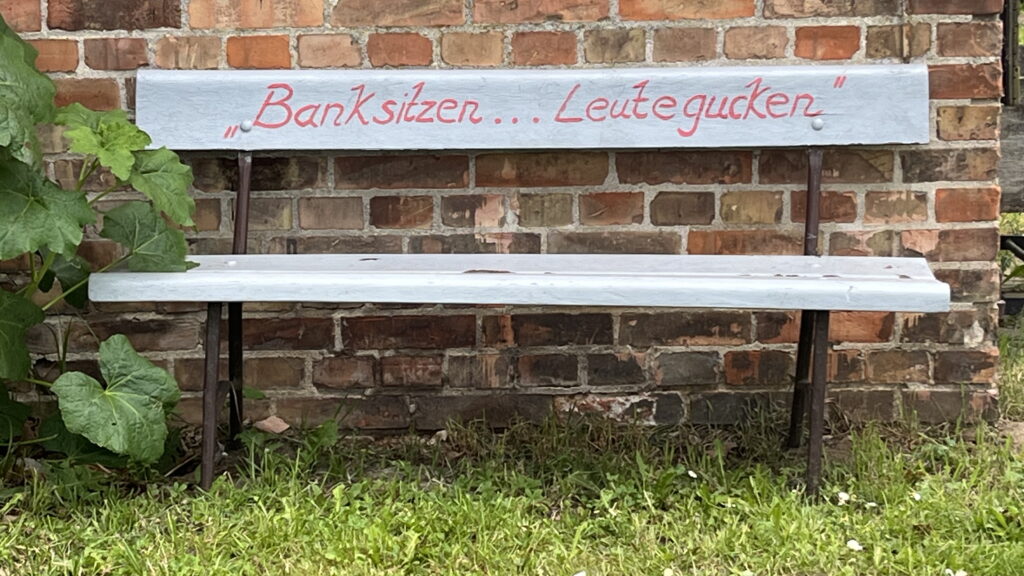 weiß gestrichene Bank mit der Aufschrift "Bank sitzen Leute gucken" vor Backsteinmauer © K. Schwahlen 2024